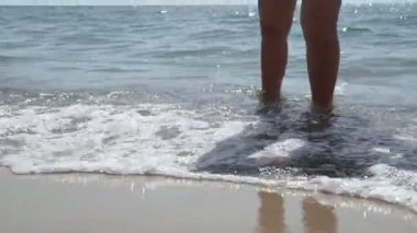 Bronzlaşmış kadın ayaklarını deniz dalgaları kaplıyor. Yaz tatili boyunca sahilde dinlenen güzel genç bir kadın. Güneşli bir günde kumsalda yatan kız bacakları. Kapat..