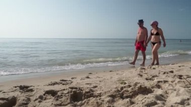 Erkek ve kadın güneşli bir günde deniz kıyısında yürüyorlar. Sıcak havalarda, turkuaz dalgalarla sulanan, kumlu deniz kıyılarında yavaşça gezinirler..