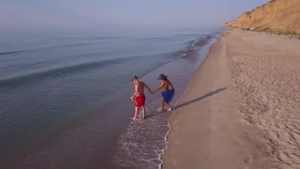 サンセットのビーチを歩く男と女ビーチで楽しい休暇を過ごしているカップルの空中撮影を追跡します ビーチで夕日の空のビーチを歩くカップル — ストック動画