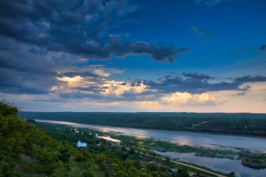 Yeşil manzaralı bir nehir ve mavi gökyüzü ile dramatik bulutlar.