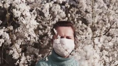 Tıbbi maskeli genç bir kadın çiçek açan bir ağaca hayran. Kadın eli, çiçek açan bir ağacın dalına dokunur. ovid-19.