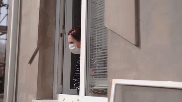顔に医療用マスクをした女性が窓から外を見ている Covid 2019 — ストック動画