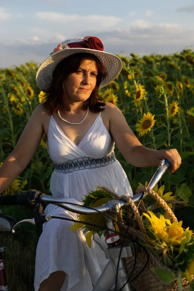 一个头戴帽子 身穿白色衣服 骑自行车的女人带着向日葵在花粉中穿行 — 图库照片
