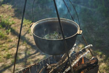 Yemek bir kazanda hazırlanır. Bovlingci kazıkta kaynıyor, arka planda ise bir çiçek tarlası var..