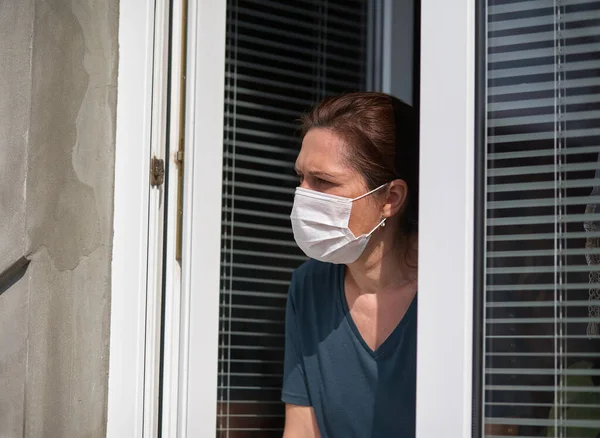 Woman in a medical face mask looks out of the window. Quarantine during the COVID-19 coronavirus epidemic
