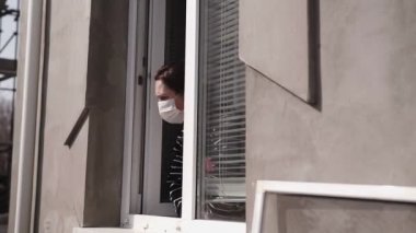 Yüzünde tıbbi maske olan bir kadın açık pencereden dışarı bakar. Covid - 2019.