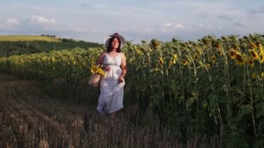 Şapkalı ve beyaz elbiseli bir kadın elinde ayçiçekleri olan bir tarlada yürüyor.