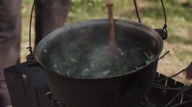 Yemek bir kazanda hazırlanır. Bovlingci kazıkta kaynıyor, arka planda ise bir çiçek tarlası var..