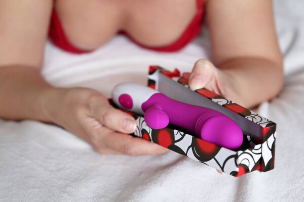 Woman with dildo in hands, sex toy for adult games. Girl in red bra laying on a bed and holding purple silicone vibrator, masturbation device