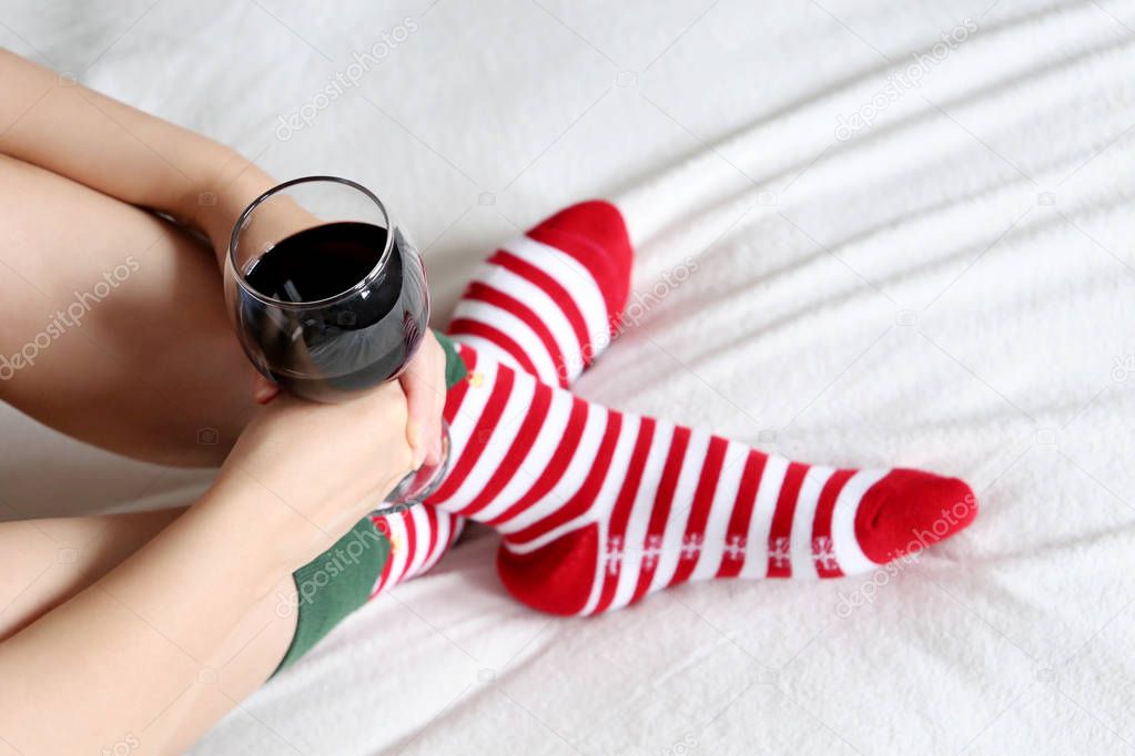 Woman drinking red wine sitting on a bed cross-legged in Christmas socks. Concept of New Year celebration, home relax and cozy atmosphere