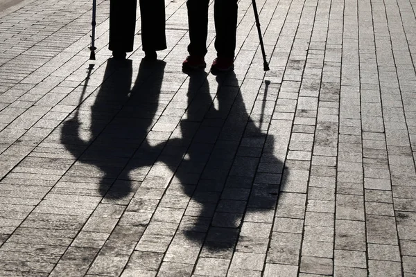 Silhouettes and shadows of two limping people walking with canes on the street. Elderly couple outdoors, concept of old age, retired or rehabilitation after injury