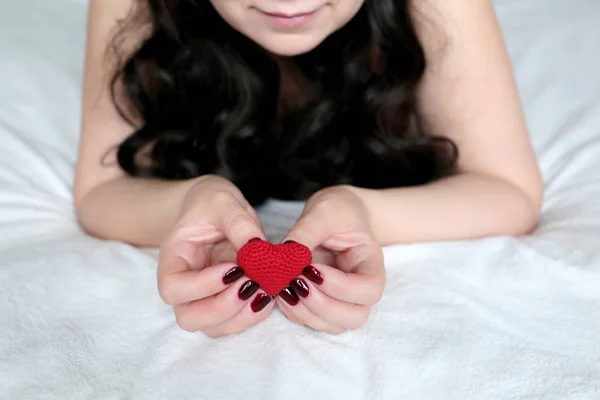 Woman in love, red knitted heart in female hands. Girl with long curly hair lying on the bed, concept of romantic date, health care, innocence or organ transplantation