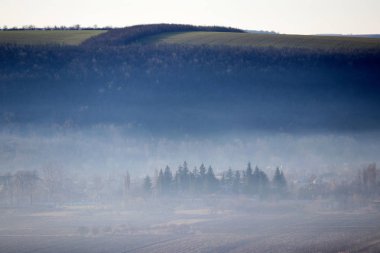 Dağlarda sis, resimli kırsal alan. Tepelerdeki tarlalar, vadideki orman ve köy sabahın erken saatlerinde