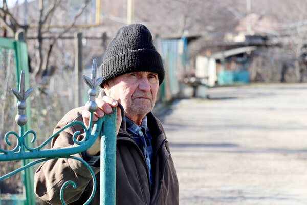 Elderly man opens the iron gate in rural yard. Unhappy face expression, concept of cold weather, life in village, old age