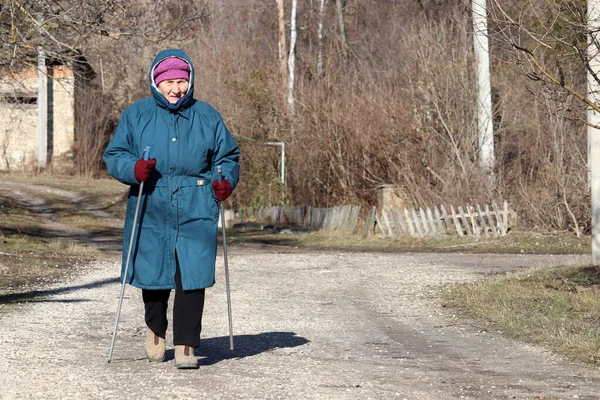 田舎道に棒で歩く高齢女性 健康な背骨や関節のためのスポーツ演習 寒い日にノルディックウォーキング 健康のための治療 — ストック写真