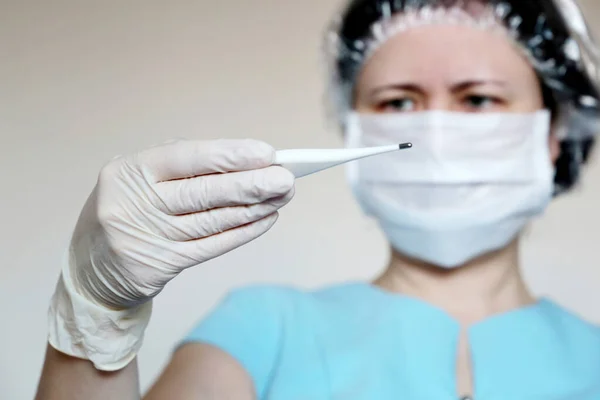 Coronavirus symptoms, woman in medical mask measures body temperature. Doctor looks at digital thermometer in her hand, concept of cold and flu