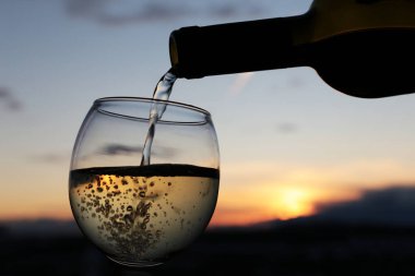 Güzel günbatımı arka planında beyaz şarap şişeden cama akıyor, turuncu güneş kara bulutların arasından parlıyor. Kutlama anlayışı, romantik akşam yemeği.