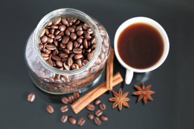 Baharatlı kahve, cam kavanozda kavrulmuş kahve çekirdekleri, yıldız anason, tarçın çubukları ve siyah cam masada beyaz bardak.