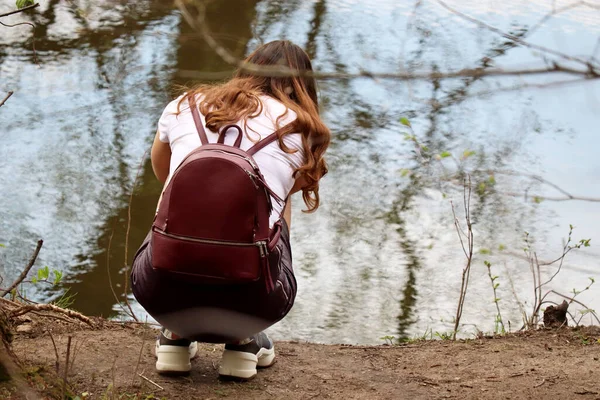 红头发姑娘背着背包蹲在河岸上 春天的户外娱乐活动 — 图库照片