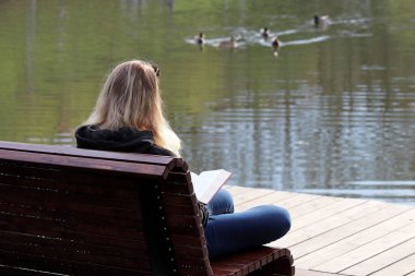 Kotlu sarışın kız, arka plandaki bir bahar parkında ahşap bankta oturuyor. Okuma kavramı, doğada rahatlık.