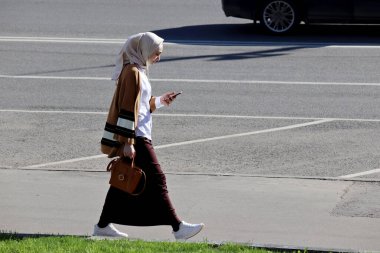 Moskova, Rusya - Mayıs 2020: Akıllı telefonları ve kulaklıkları olan genç Müslüman kadın şehir caddesinde yürüyor. Kaldırımda tesettürlü güzel kız