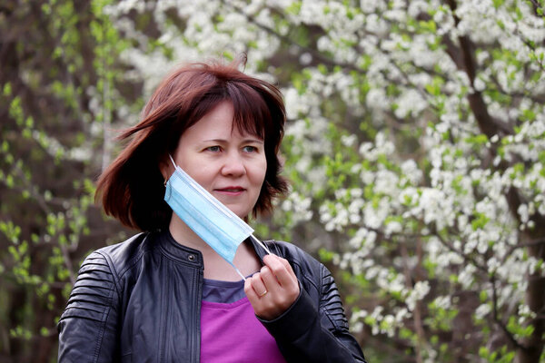 Joyful woman removing medical protective mask in a spring garden on cherry blossom background. Concept of enjoying the flowers smell, fresh air, end of the quarantine during covid-19 coronavirus pande