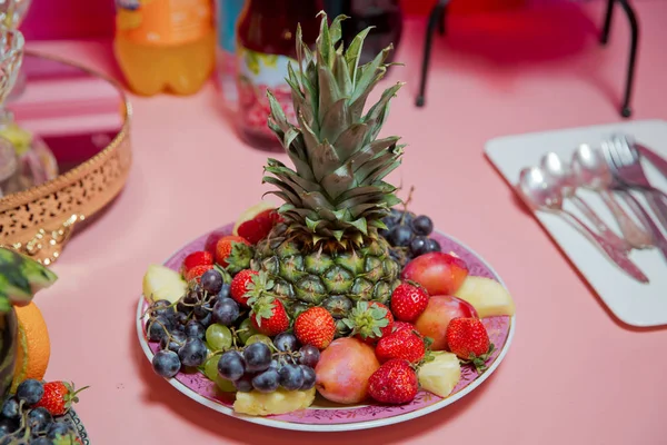 pink plate, pineapple, strawberry, white and black grapes, red cherry, chopped apples. pink background.