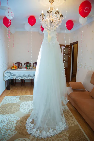 White wedding dress hangs on a chandelier in room .The perfect wedding dress in the room of the bride . Wedding detail photography. Beautiful white wedding dress. Bridal morning