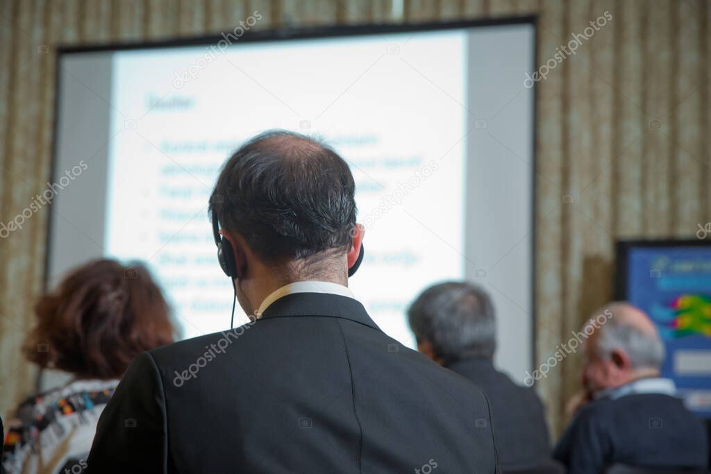 Man with headset at the conference . The audience wearing head phone for online translation . Unrecognizable people using in ear headphones for translation during event