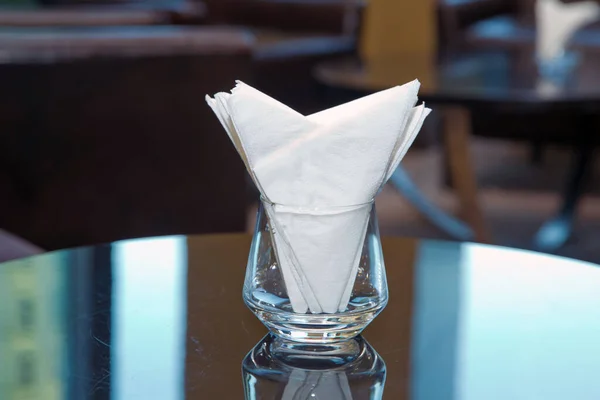 Θαμπάδα εικόνας-λευκή χαρτοπετσέτα τοποθετείται σε ένα ποτήρι στο τραπέζι στην αίθουσα δεξιώσεων. Τραπέζι φαγητού με διακοσμητική χαρτοπετσέτα, γυάλινα επιτραπέζια σκεύη στην τραπεζαρία του ξενοδοχείου . — Φωτογραφία Αρχείου