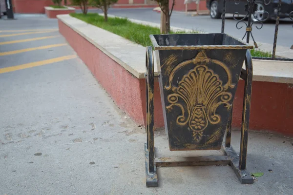 Black trash can in the yard of the building . Black copper metal garbage bin in building with blurred . Garbage can on top of asphalt .