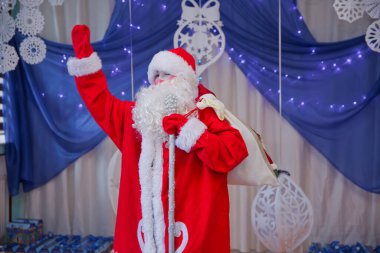 Peder Frost mikrofonla konuşuyor. Noel Baba aleyhinde Noel şarkıları söylüyor. Noel Baba kostümlü bir adam mikrofonla poz veriyor..