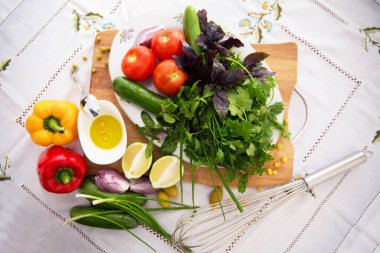 Canlı domatesler, salatalıklar, doğranmış kırmızı soğanlar, yeşiller, fesleğenler, ahşap bir tahtada kişniş. Küçük beyaz bir kasede zeytin. Doğranmış yeşil limon. Soğan. Yumurta çırpmak için metal çırpma.