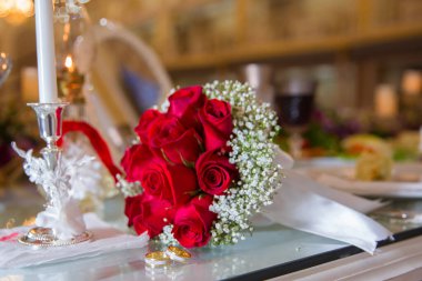 Kızıl düğün çiçekleri. Nişan yüzükleriyle gelin ve damat masaya konmuş ve yanlarında bir düğün buketi var. Beyaz masa. Çiçek buketinin yanında alyans seçici bir odak noktası