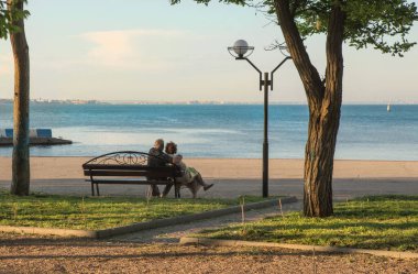 Yaşlı evli çift, deniz kenarındaki bir bankta kucaklaşarak otururlar.