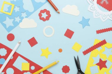 mavi bir arka plan üzerinde dağınık renkli kağıt kalem, makas ve basit geometrik şekiller kesilmiş