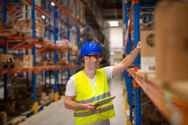 Warehouse worker looking at shelves with packages and checking inventory of large warehouse storage distribution area.