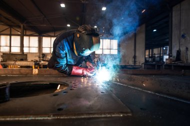 Professional industrial welder welding metal parts in metalworking factory. Worker wearing protective mask and gloves. Flash and sparks flying.