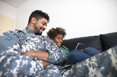 Soldier in military uniform being home with family enjoying and watching cartoons with his daughter on tablet computer.