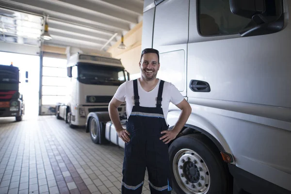 Portrait of positive smiling truck serviceman standing by truck vehicle in workshop. Truck vehicle maintenance and servicing.