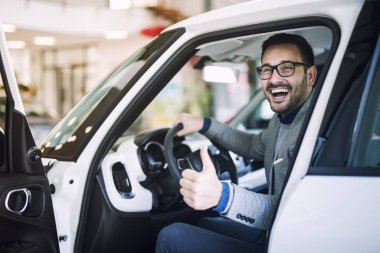 Happy satisfied customer just bought a brand new car at vehicle dealership. Car buyer sitting at drivers position and holding thumbs up.