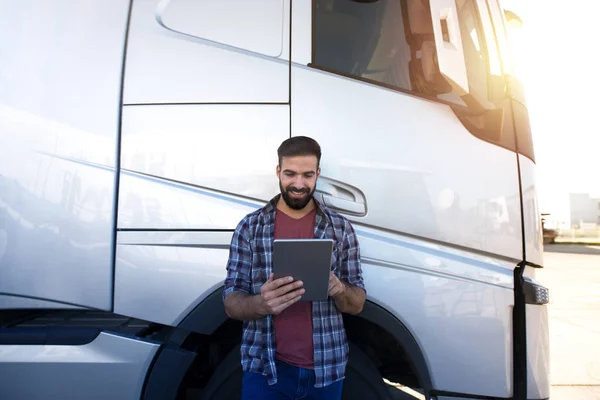 Professional truck driver. Young bearded trucker checking his route on tablet computer and standing by long vehicle. Transportation service.