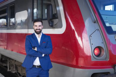 Mavi üniformalı genç sakallı tren şoförünün portresi modern metro treninin önünde gururla duruyor. Ulaşım mesleği ve hizmet.