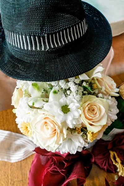 wedding bouquet of beige roses and chamomiles under a black male hat