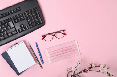 Tıbbi maskeli, klavyeli, not defterli ve pembe arka planda gözlüklü çiçek açan dallar. Coronavirus COVID-19