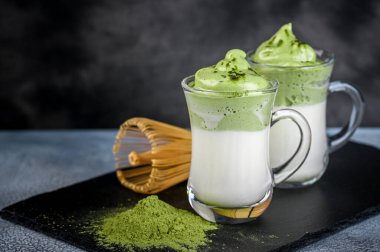 İki fincan Dalgona Matcha Latte, koyu arkaplanda kremalı bir kibrit. Kibrit yeşil çay.
