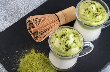 İki fincan Dalgona Matcha Latte, koyu arkaplanda kremalı bir kibrit. Kibrit yeşil çay.