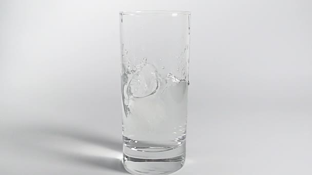 Dos trozos de hielo cayendo en un vaso transparente de agua clara uno tras otro. Movimiento lento sobre fondo blanco con sombra . — Vídeos de Stock