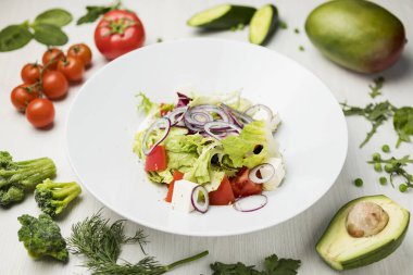 Taze sebze salatası salatası büyük beyaz bir tabakta domates, avokado, maydanoz, salatalık, bezelye, brokoli karışımı beyaz bir masada.