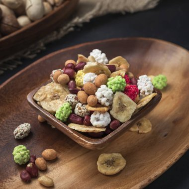 Kare bir ahşap kaseye yerleştirilmiş fındık ve meyve karışımı ve koyu arka planda oval ahşap tepsi ve çuval bezi.